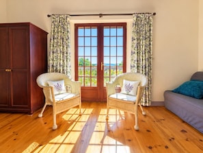 Une pièce avec un plancher en bois, deux fauteuils en osier avec des coussins, un canapé, une armoire en bois et une grande fenêtre avec des rideaux.