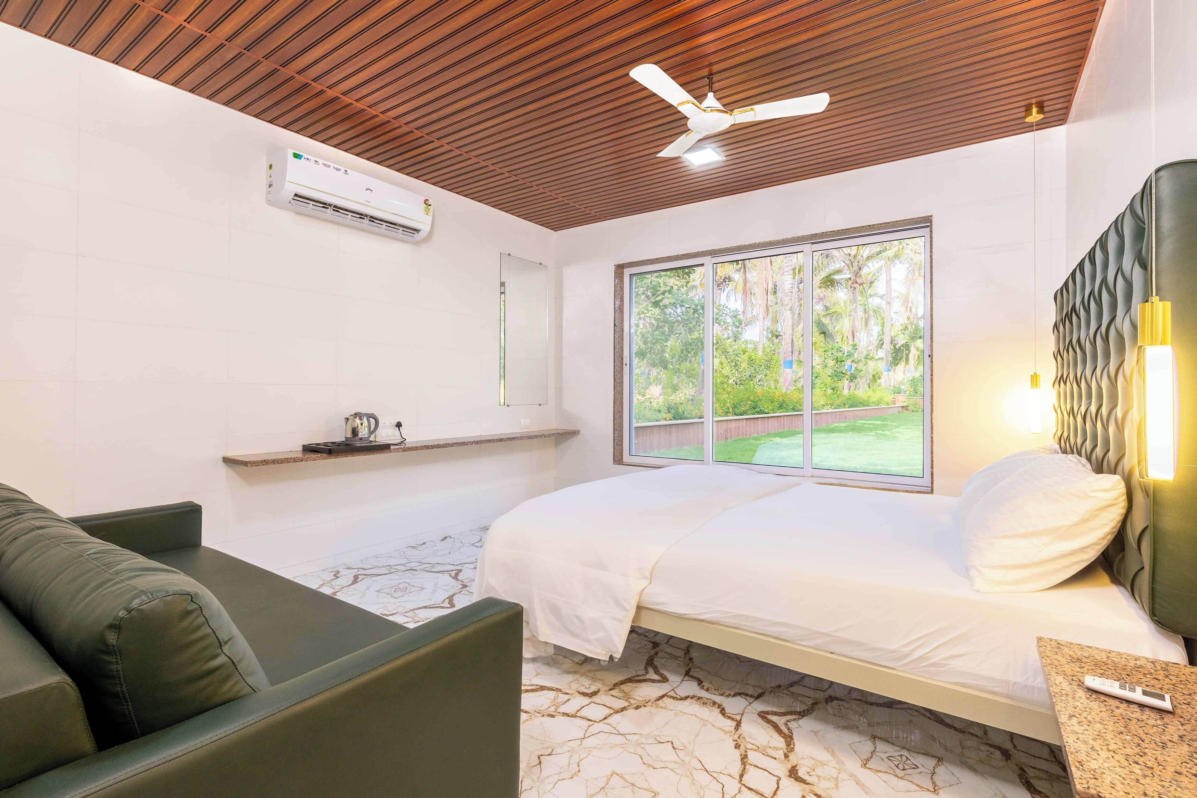 A modern bedroom with a bed, a green sofa, a ceiling fan, and a window with a view of greenery.