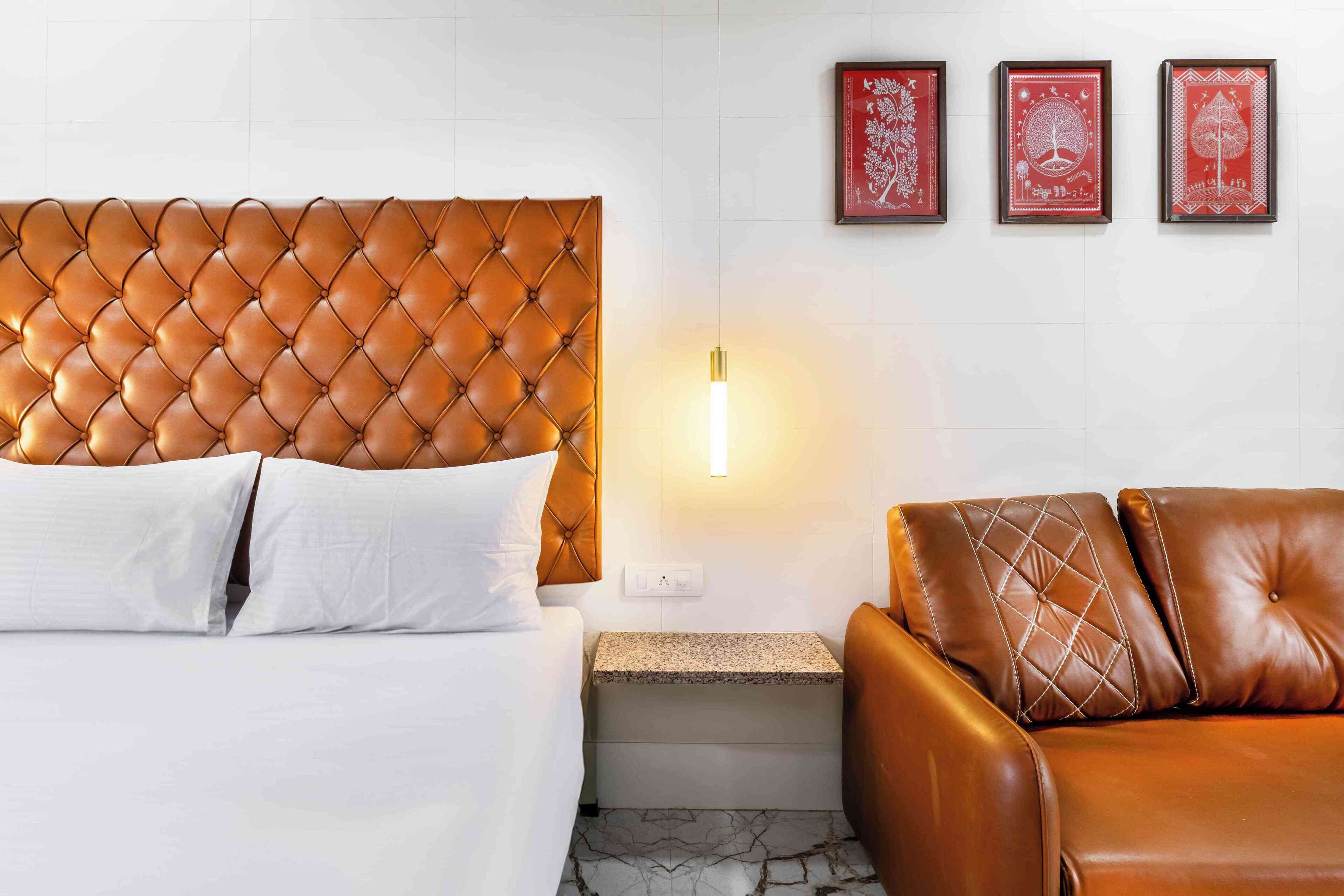Une chambre d’hôtel avec une tête de lit capitonnée, un lit avec du linge de maison blanc, un canapé en cuir marron, une table de chevet avec une lampe et trois œuvres d’art encadrées accrochées au mur.