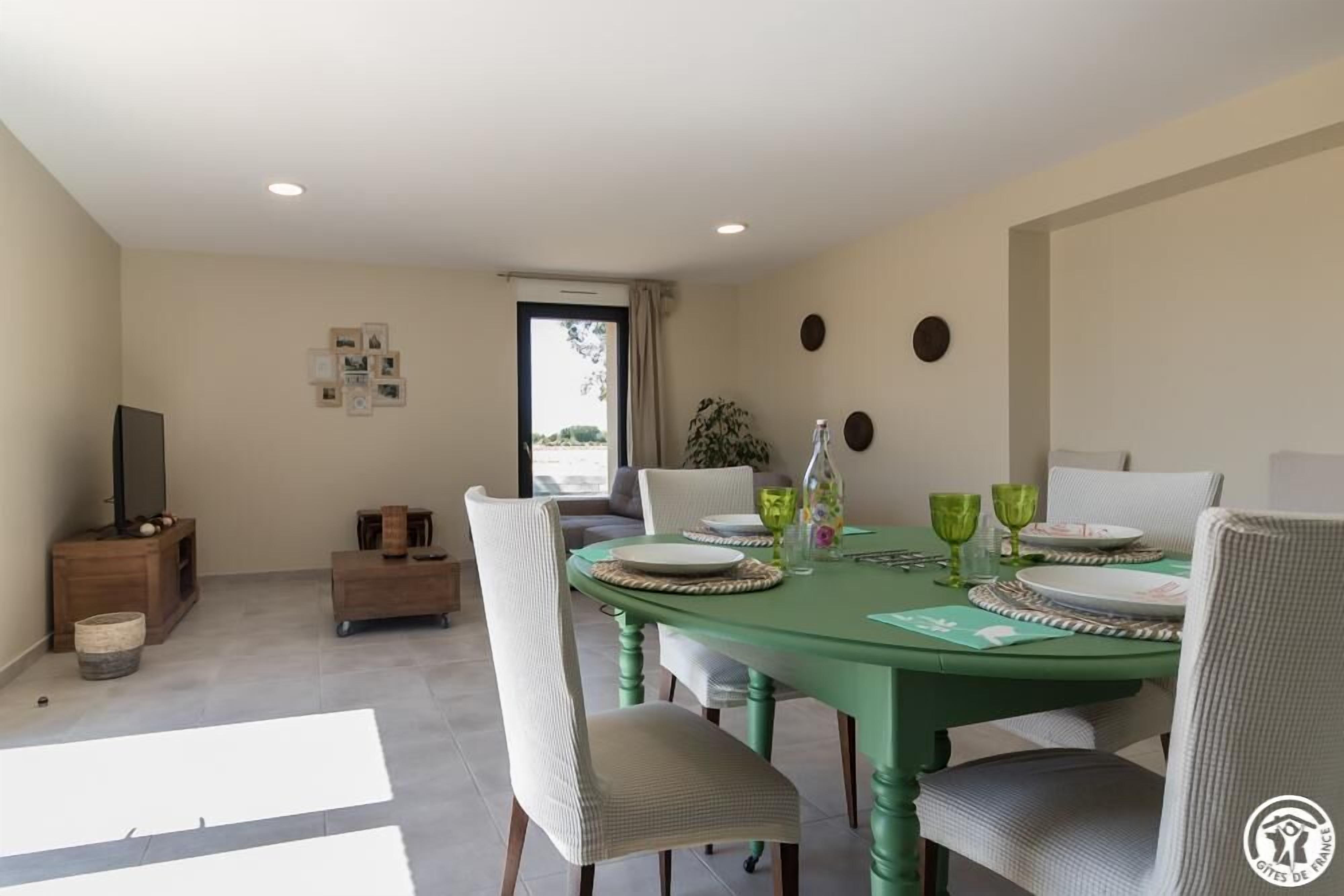 Een eethoek met een groene tafel voor vier personen, omringd door witte stoelen. Links staat een houten kast met een televisie en er is een plant bij het raam.