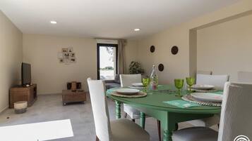 Een eethoek met een groene tafel voor vier personen, omringd door witte stoelen. Links staat een houten kast met een televisie en er is een plant bij het raam.
