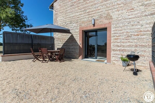 An outdoor patio with a table and chairs, a barbecue grill, and a building with a brick facade.