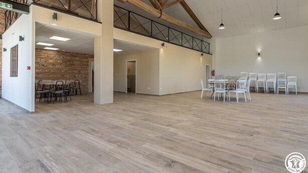 Un vaste espace intérieur doté d’un haut plafond, de poutres en bois et d’une mezzanine. On y trouve plusieurs tables et chaises disposées pour les repas ou les réunions.