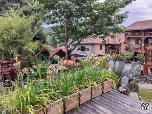 Un jardí amb diverses plantes i flors, un pont de fusta i un edifici amb teulada de teules.
