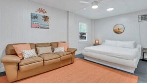 A bedroom with a white bed, a brown sofa, a white wall with a picture, and a ceiling fan. - 3-Bed, 1-Bath Comfort & Style
 (Tybee Island)