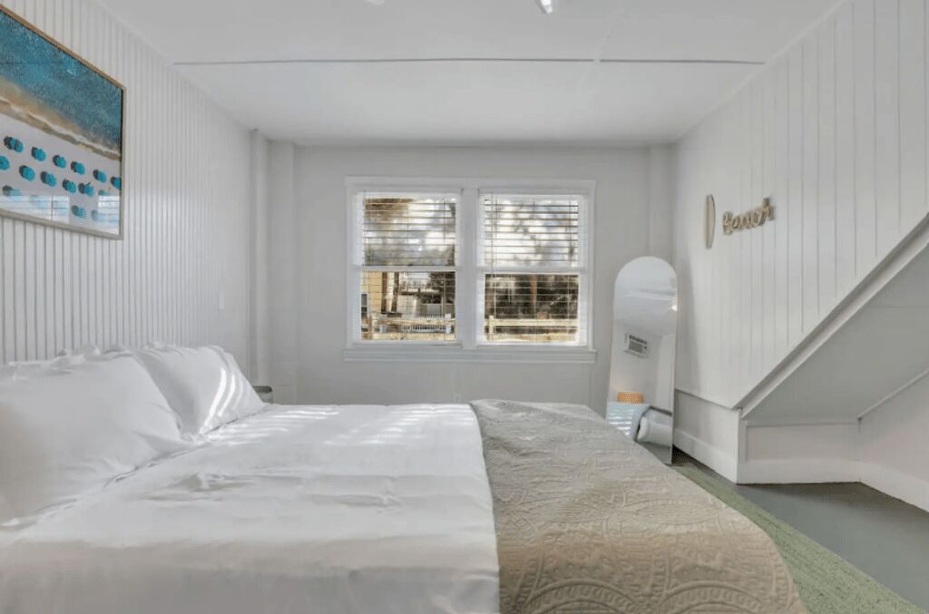 Une chambre à coucher avec un lit, une fenêtre avec des stores, un tableau au mur et un escalier.