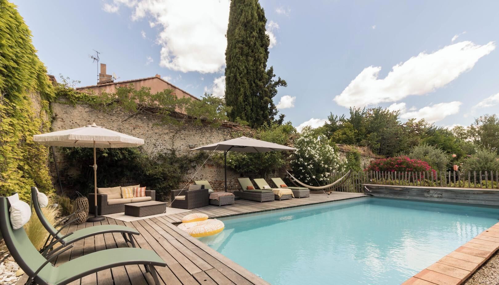 Ein Schwimmbecken mit Liegestühlen und Sonnenschirmen, umgeben von einer Holzterrasse und viel Grün.
