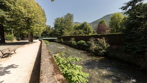 A park with a paved walkway, a bench, and a clear stream. - Vacations Cures Luchon (Bagnères-de-Luchon)