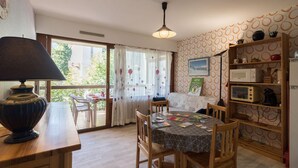 A cozy room with a wooden table, chairs, a lamp, a bed, a shelf with kitchen appliances, and a window with curtains. - Vacations Cures Luchon (Bagnères-de-Luchon)