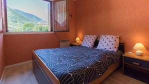 A bedroom with a bed, two bedside tables, a window with curtains, and a view of greenery outside. - Vacations Cures Luchon (Bagnères-de-Luchon)