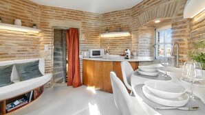 A compact kitchen with a brick wall, a white countertop, and a dining area with white chairs and plates.