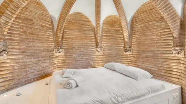 A bedroom with a brick wall and arched ceiling, a bed with white linens, and a small bedside table.