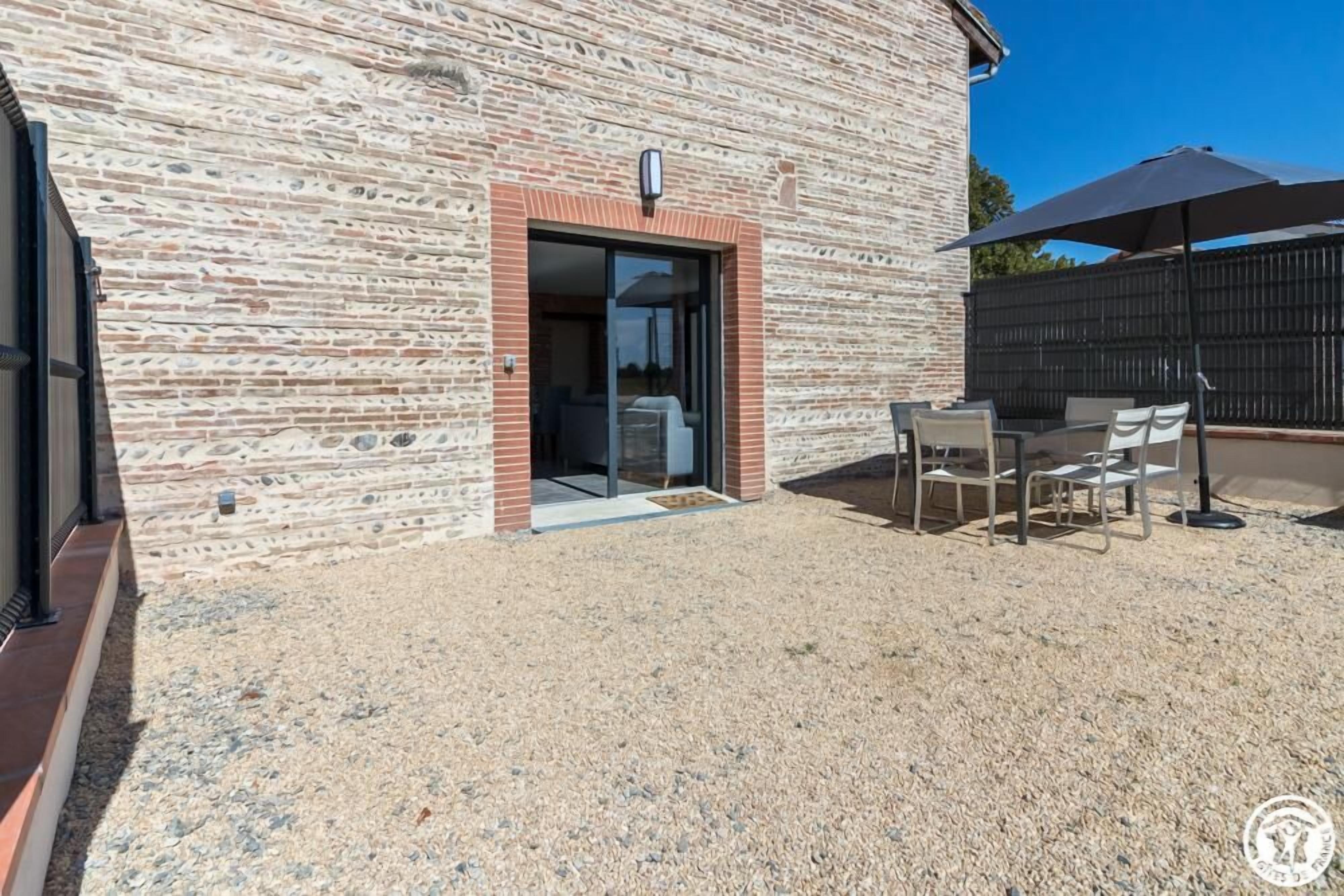Un edificio con fachada de piedra, puerta de acceso de vidrio y un patio con mobiliario exterior.