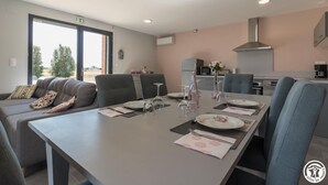 A modern dining area with a long table set for four, a sofa with cushions, and a kitchen in the background. - Domaine le Caprougeat - Chic countryside (Daux)