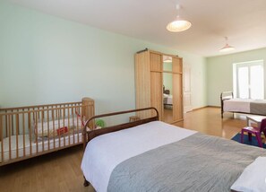 Een kamer met een groot bed, een babybedje, een bureau en een kledingkast.