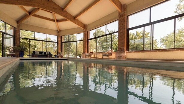 Une piscine intérieure avec des parois en verre transparent, des poutres en bois et des espaces aménagés pour s’asseoir.