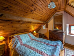 A bedroom with a wooden ceiling, a bed with a striped blanket, a nightstand with a lamp, a dresser, and a window with a view.