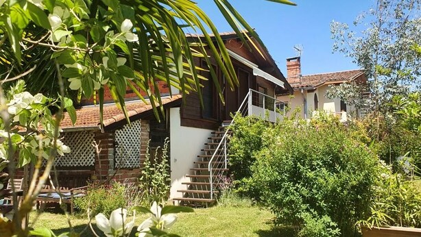 Une maison à deux étages, avec un escalier apparent menant à l’étage supérieur, entourée d’une végétation luxuriante et de plantes en fleurs.