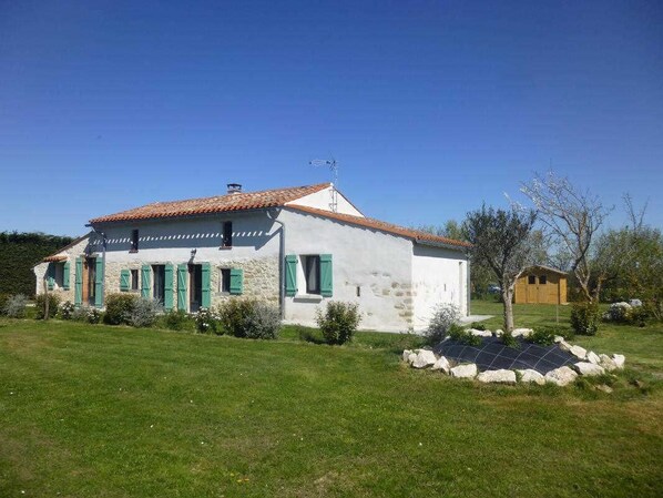 Un bâtiment blanc, doté d’un toit en tuiles et de volets verts, entouré d’une pelouse bien entretenue et d’un ensemble de panneaux solaires.