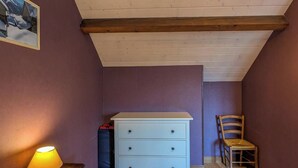 Une pièce avec un plafond en pente, une commode blanche à tiroirs, une chaise en bois et une lampe sur une table.