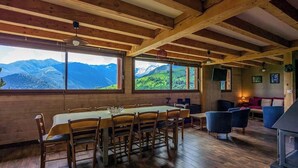 Un espace repas comprenant une table et des chaises, de grandes fenêtres offrant une vue sur les montagnes, et un coin salon confortable avec un canapé et des fauteuils.