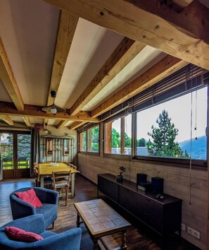 Une pièce avec un plafond en bois, un coin repas, un canapé, un meuble pour la télévision et de grandes fenêtres offrant une vue sur les arbres et les montagnes.