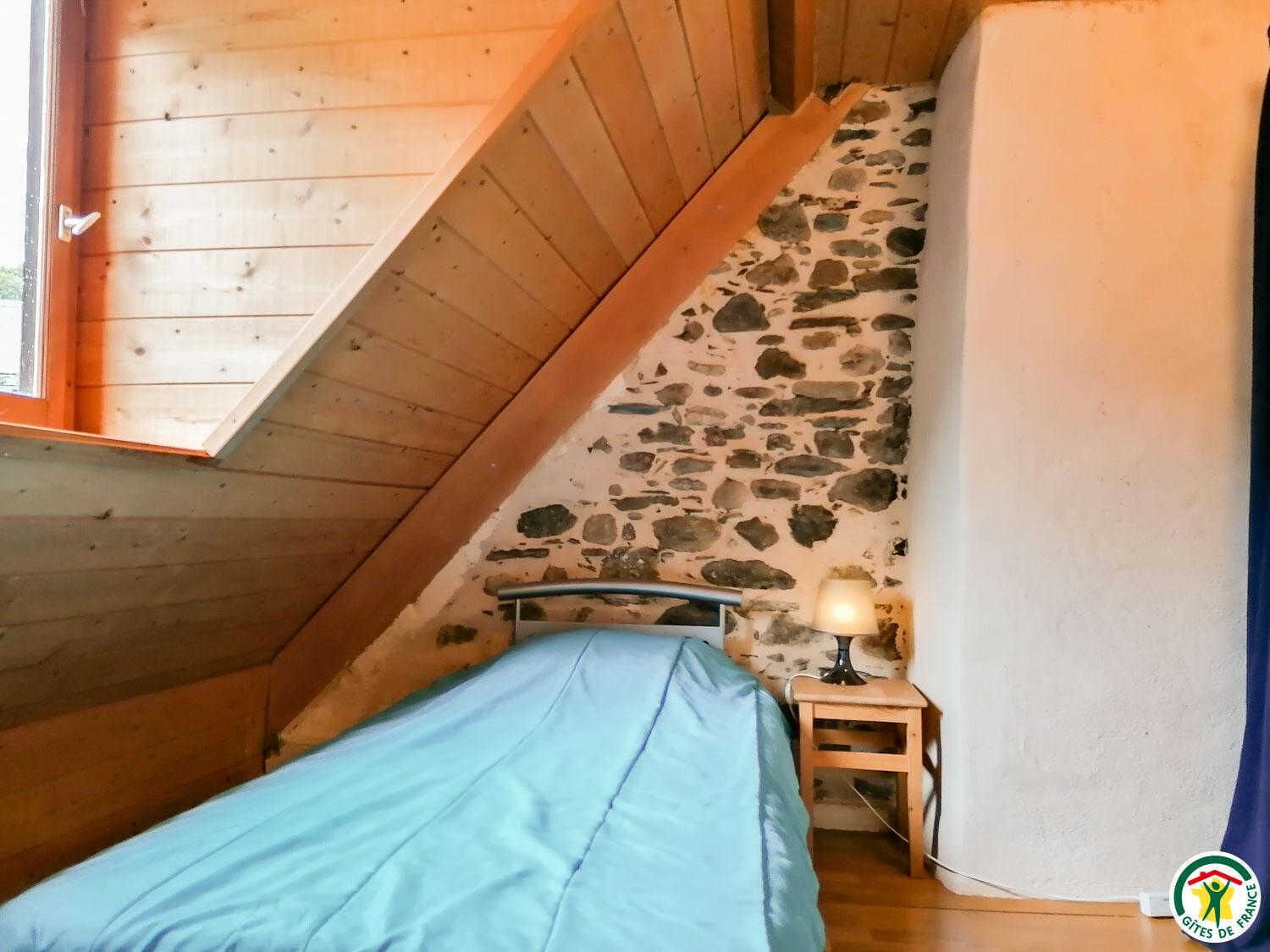 A bedroom with a sloped ceiling, a bed, a nightstand, a lamp, and a stone wall.