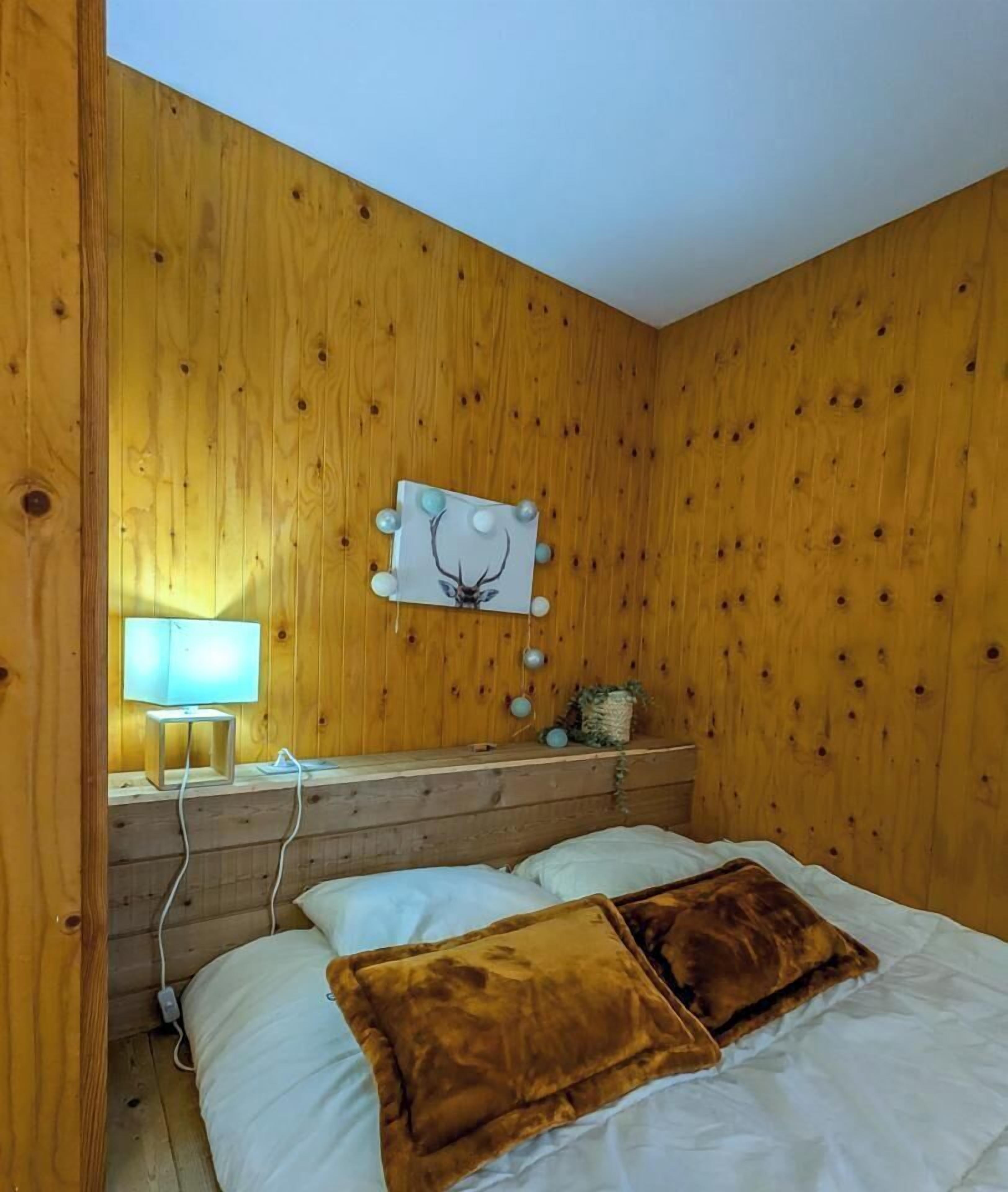 A wooden-paneled room with a bed, a bedside lamp, a wall-mounted decoration, and a small shelf with a plant.