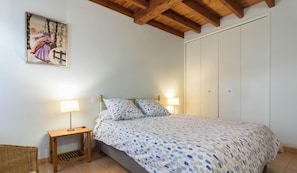Un dormitori amb un llit, làmpades de nit, una petita taula de nit de fusta, una cadira de vimbles i un quadre penjat a la paret.