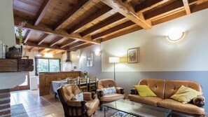 Una sala de estar acogedora con un sofá de cuero, sillones, una mesa de centro de vidrio, vigas de madera en el techo y un comedor con mesas y sillas de madera.