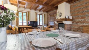 Un coin repas avec une nappe à carreaux, des chaises en bois, une cheminée et un téléviseur en arrière-plan.