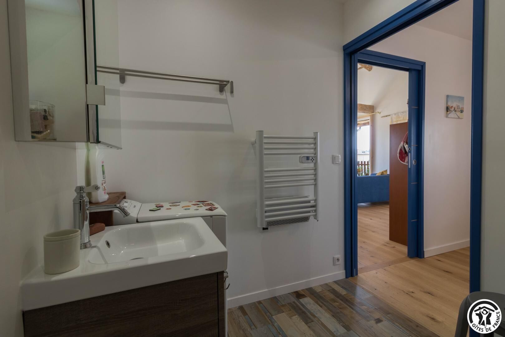 Une salle de bain avec un lavabo blanc, un miroir et un porte-serviettes. Le sol est en bois et une porte bleue donne accès à une autre pièce.