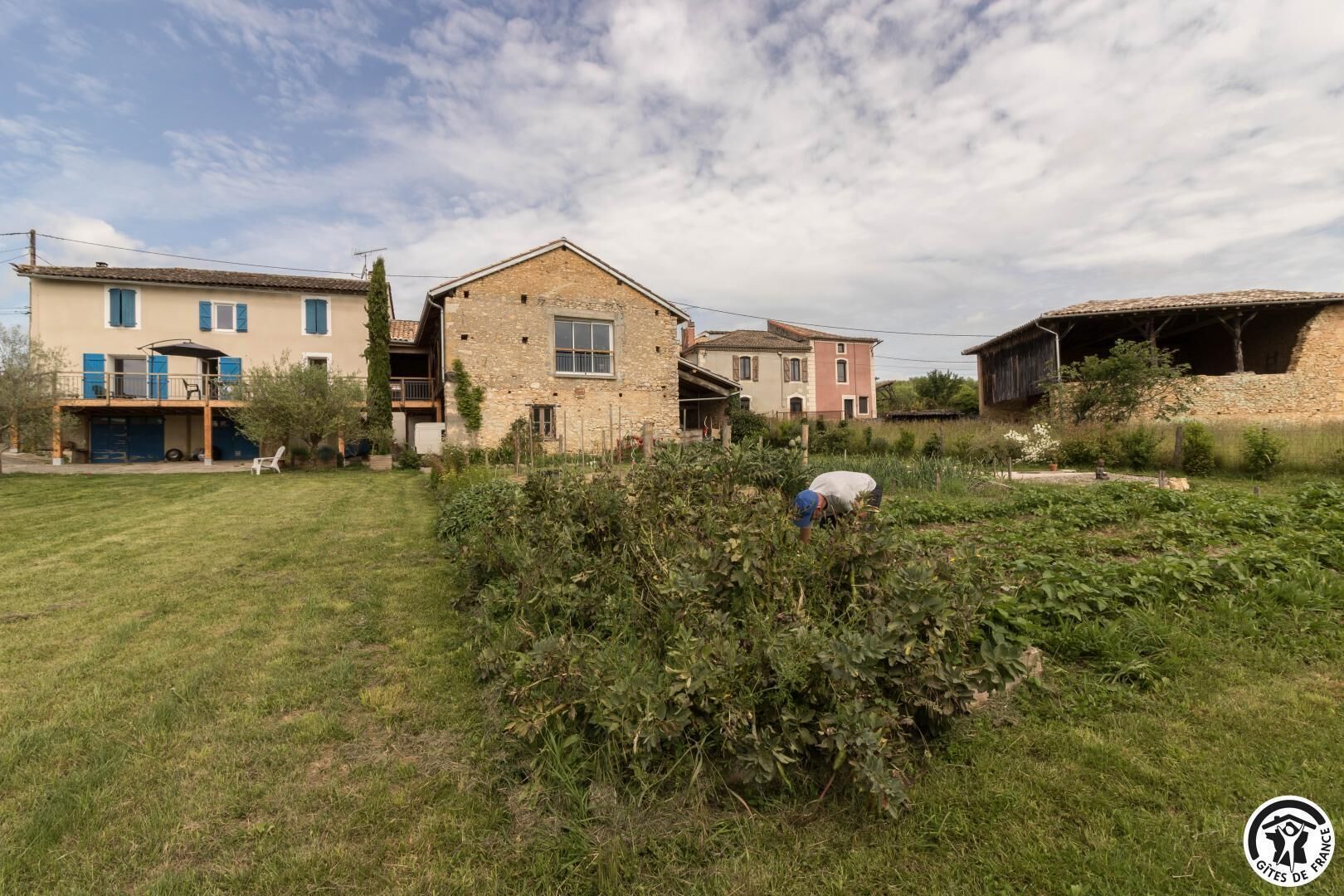 Een landelijk gelegen pand met een stenen huis, een gazon en een persoon die planten verzorgt.
