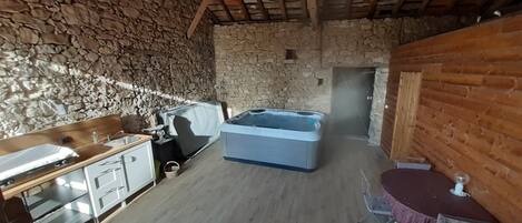 A rustic cabin with a stone wall, wooden ceiling beams, a hot tub, a kitchenette, and a dining area.