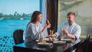 A dining table with plates of food and drinks, a view of the sea, and a window with curtains. - Le Fayan Nile Cruise - Mondays from Aswan & Thursdays from Luxor (Luxor)