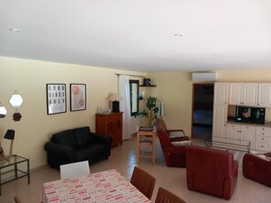 Ett vardagsrum med en svart soffa, en röd fåtölj, ett matbord med ett mönstrat bordstycke och en köksdel med vita skåp.
