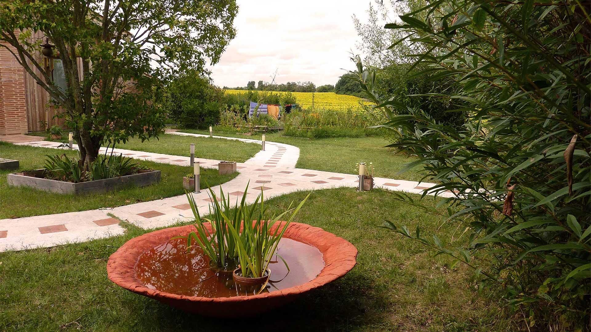 Un jardín con un sendero empedrado, una jardinera grande con plantas y un campo de flores amarillas al fondo.