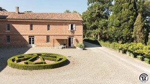 Un grand bâtiment en briques surmonté d’un toit de tuiles, un labyrinthe circulaire formé d’arbustes au premier plan et une rangée de plantes en pot le long de l’allée.