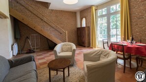 Un vaste salon agrémenté d’un grand escalier en bois, un coin repas avec une nappe rouge et une vue sur l’extérieur grâce à de grandes fenêtres.