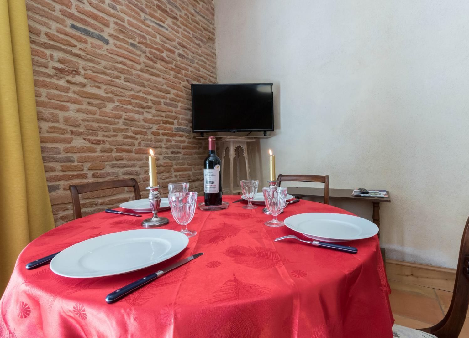 A dining table set for four with a red tablecloth, wine glasses, plates, and cutlery. A bottle of wine and two candles are on the table.