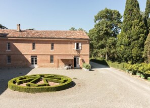 Un grand bâtiment en briques surmonté d’un toit de tuiles, un labyrinthe circulaire formé d’arbustes au premier plan et une rangée de plantes en pot le long de l’allée.