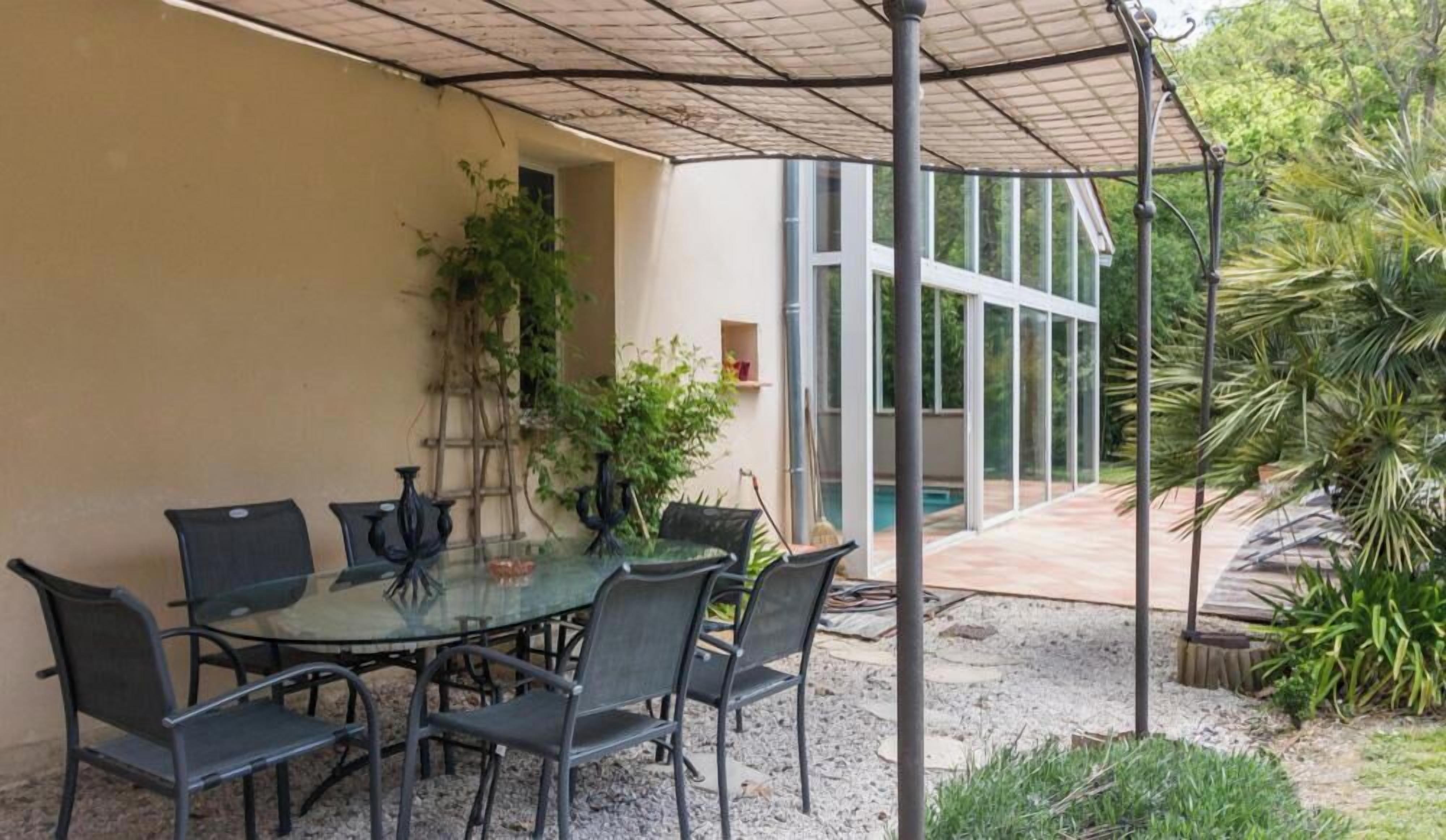A covered outdoor dining area with a glass table and metal chairs, surrounded by plants and a view of a swimming pool.