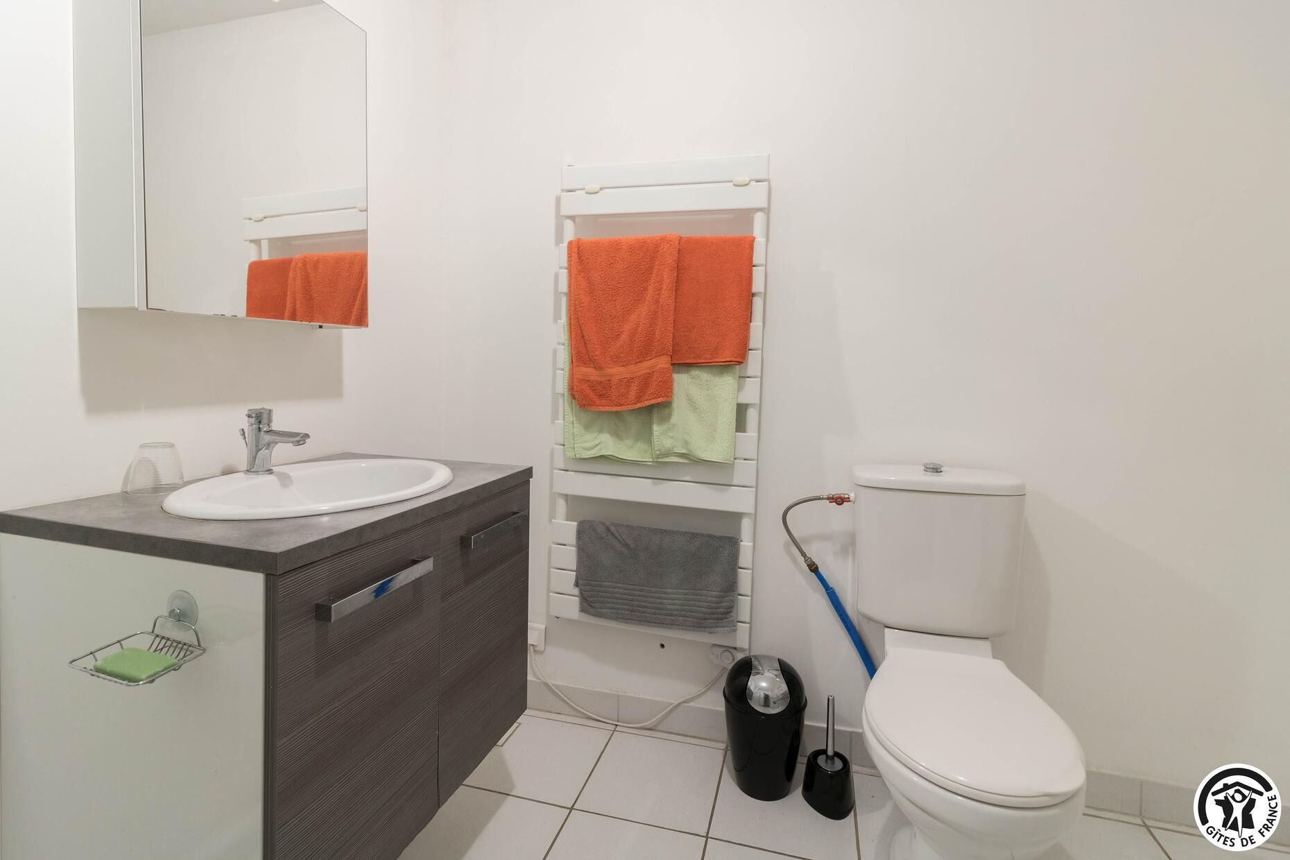 Ein Badezimmer mit weißem Waschbecken, Spiegel und Toilette. Handtücher hängen an einem Wäscheständer, und eine Mülltonne steht neben der Toilette.