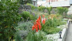 Un jardin avec différentes plantes et fleurs, un mur de pierre et, au fond, un bâtiment doté d’un toit en pente.