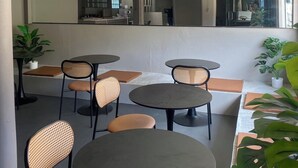 Un área de estar interior moderna con mesas redondas y sillas de mimbre, que incluye un espejo grande y plantas en macetas.