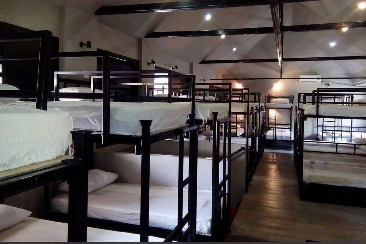 A dormitory-style room with bunk beds arranged in rows, wooden railings, and a high ceiling with exposed beams.