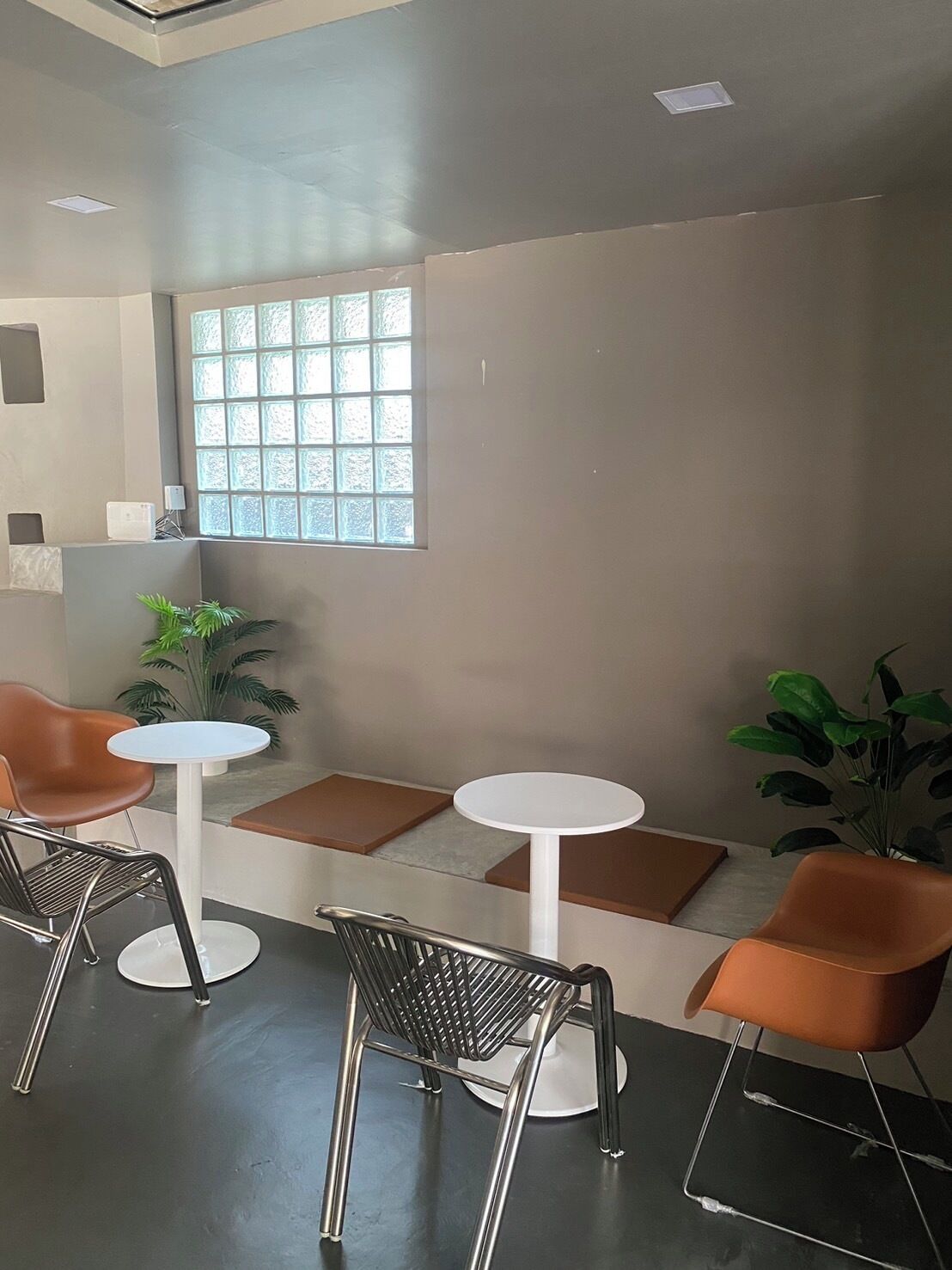 Un interior moderno con dos mesas redondas y cuatro sillas, un amplio ventanal con vistas y una planta en la esquina.