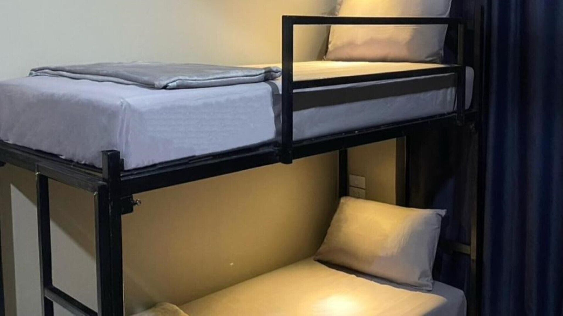 Uma cama beliche com colchão e almofadas, uma mesinha de cabeceira e uma cortina.