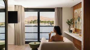 Habitación con un ventanal grande que da a un cuerpo de agua y montañas, un televisor de pantalla plana en la pared, y una pequeña mesa redonda con un florero y fruta.