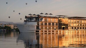 Un gran barco de recreo fluvial con múltiples cubiertas y zonas de estar al aire libre, reflejado en el agua.
